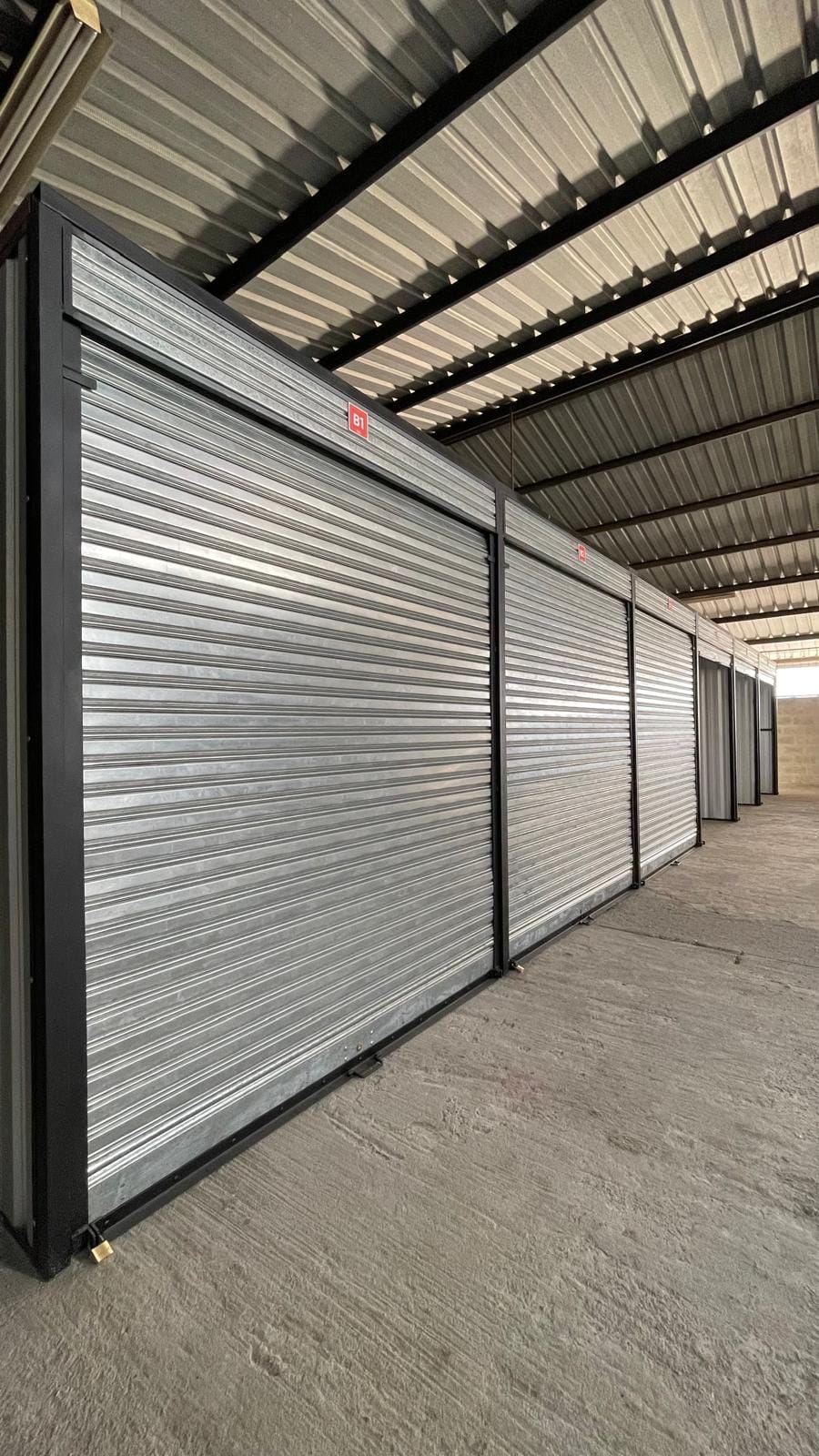 Pasillo de bodegas con las puertas metálicas galvanizadas cerradas, marcos negros, etiquetas rojas de identificación y techo de lámina.